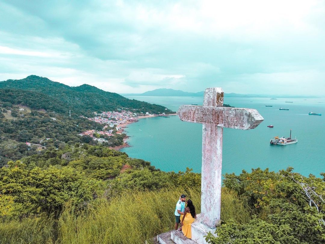 Cruz de concreto en lo alto del cerro, con la bahía turquesa, el pueblo y los barcos al fondo